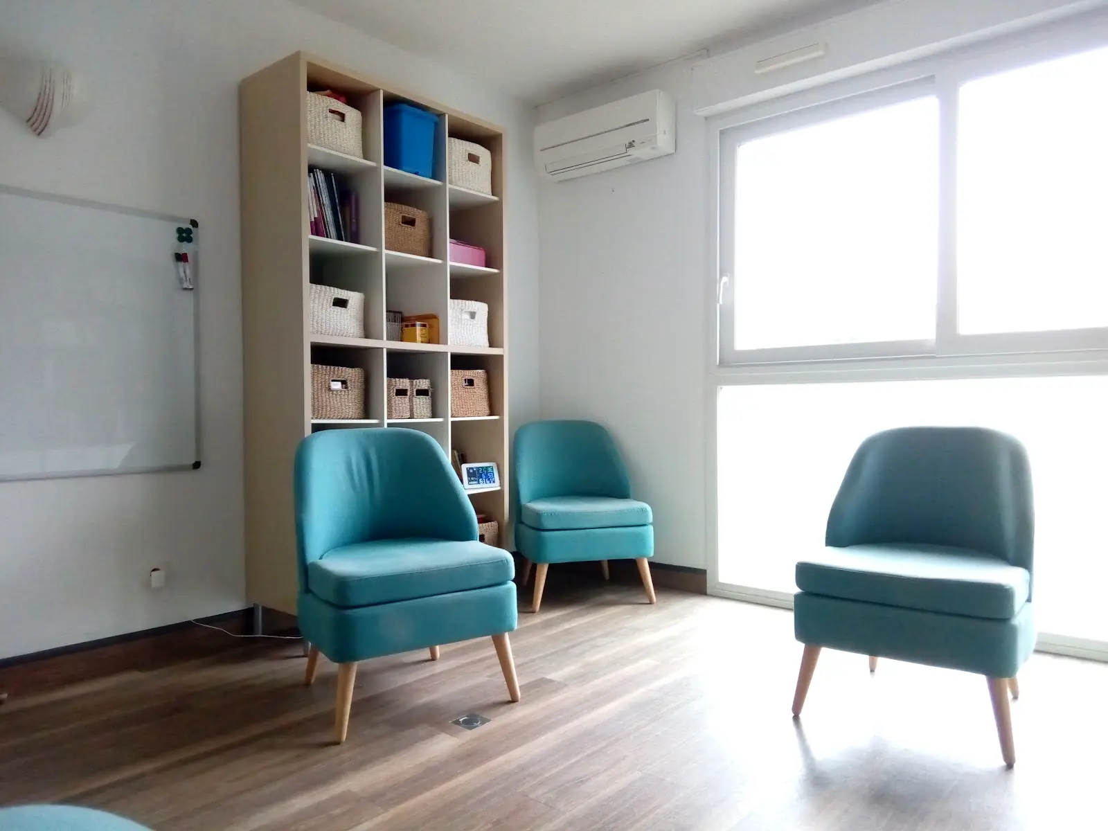 La salle de consultation : trois fauteuils bleu canard, une bibliothèque en bois clair et une grande fenêtre
